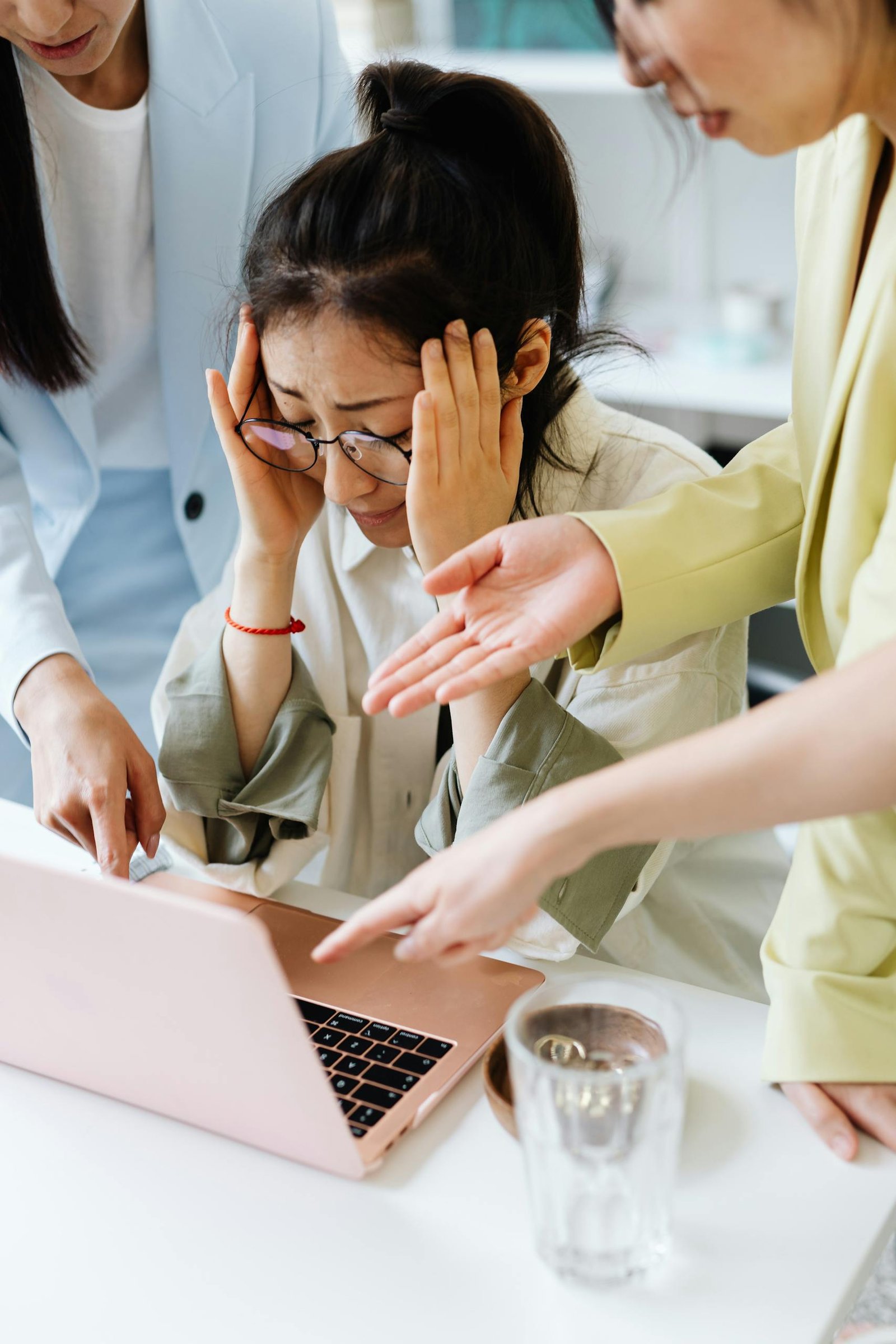 An adult woman looking stressed at work, surrounded by coworkers pointing at a laptop indoors.