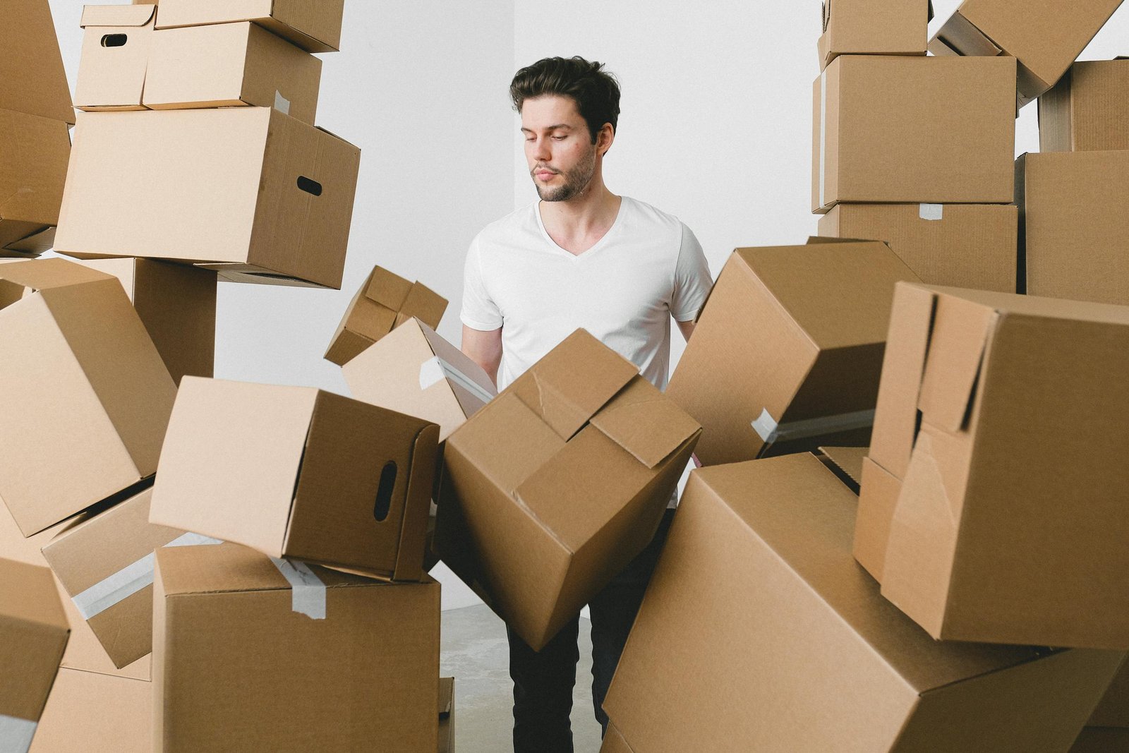 Bearded male looking down among piles of carton containers after moving into new house on light background